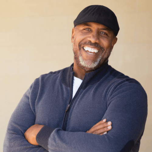 Smiling bearded man in a black cap and navy zip sweater with his arms crossed